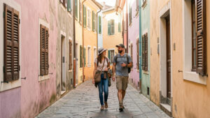 Une ruelle historique européenne bordée de façades anciennes colorées, où deux voyageurs admirent l’architecture locale dans la douce lumière du matin.